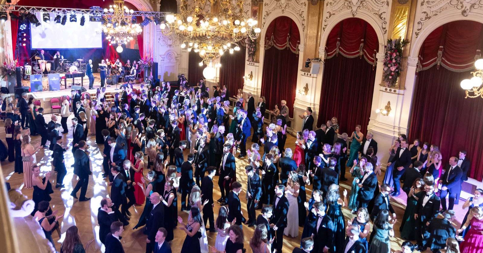 Großer Ballsaal im Palais Kaufmännischer Verein Linz, dicht gefüllt mit elegant gekleideten Gästen, die zur Live-Musik einer Band auf der Bühne tanzen.