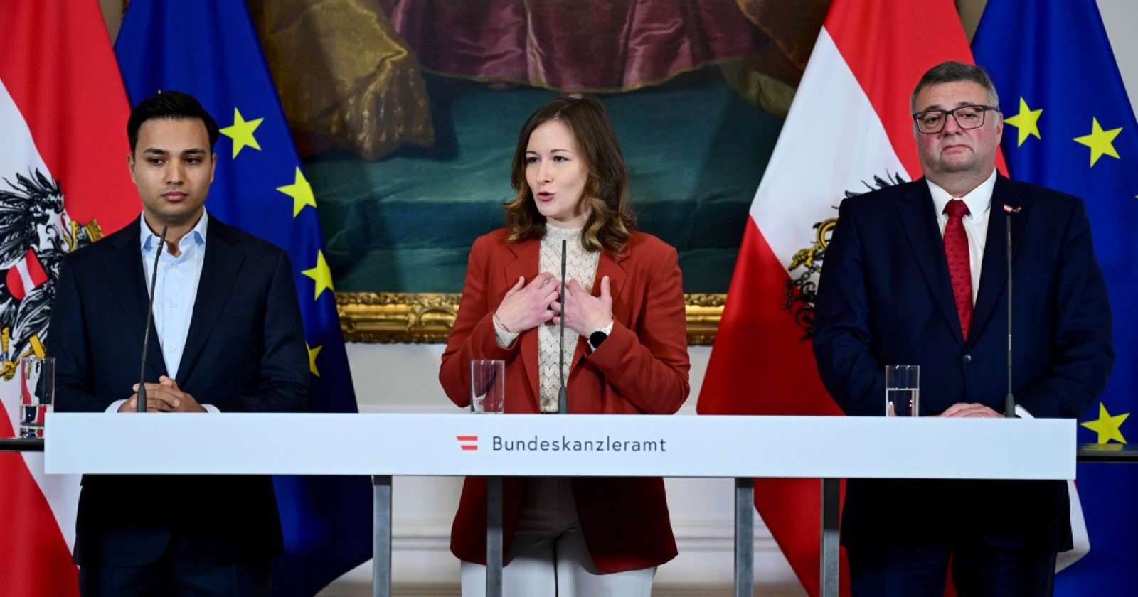 Drei Personen bei einer Pressekonferenz im Bundeskanzleramt: eine Frau in der Mitte spricht gestikulierend, flankiert von zwei Männern. Hinter ihnen stehen Österreich- und EU-Fahnen.