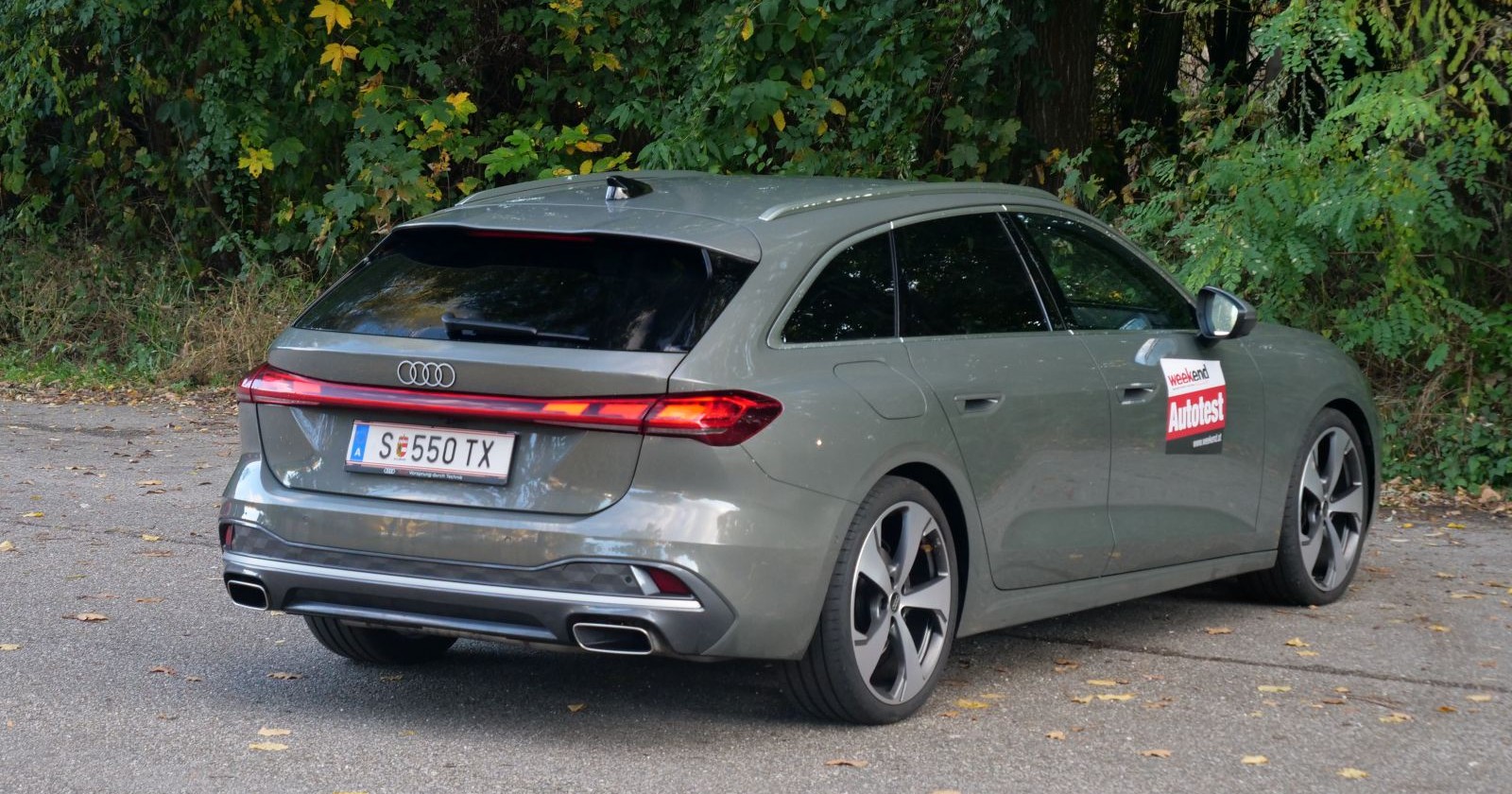 Ein grauer Audi A5 von hinten. Das Auto parkt am Rande eines Laubwaldes. Am Boden sind einige Blätter zu sehen, die herbstlich verfärbt sind.