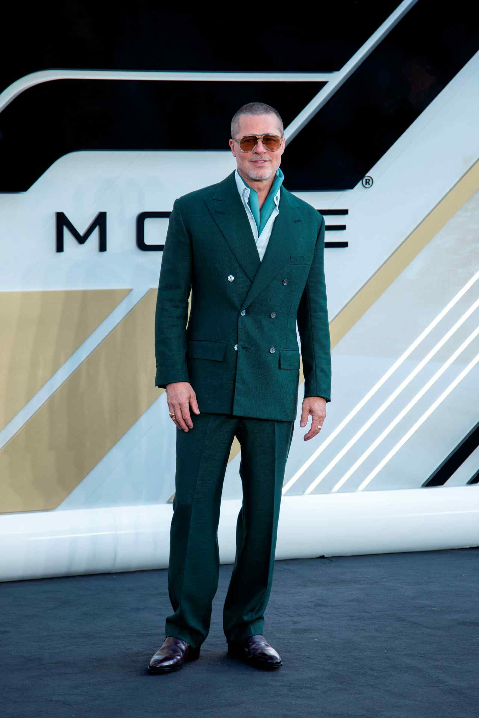 Ein Mann steht vor einer modernen, grafisch gestalteten Rückwand und trägt einen dunkelgrünen Anzug mit offenem Hemd und Sonnenbrille.