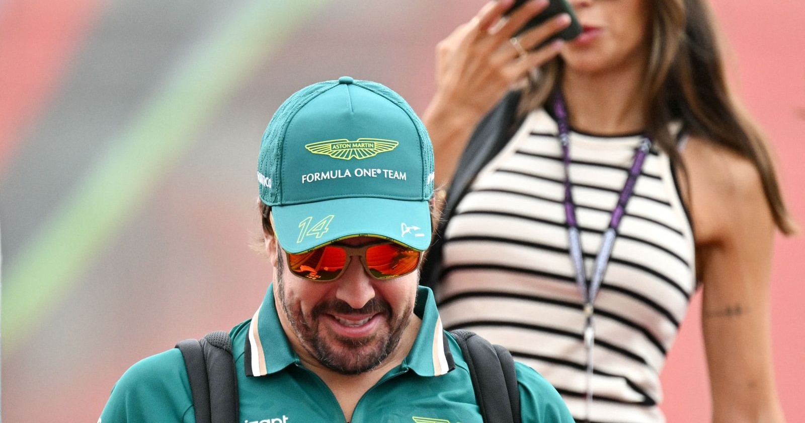 Fernando Alonso in Aston-Martin-Teamkleidung mit Kappe und Sonnenbrille im Paddock, im Hintergrund Melissa Jiménez mit gestreiftem Top und Handy am Ohr. Die beiden erwarten ihr erstes gemeinsames Kind.