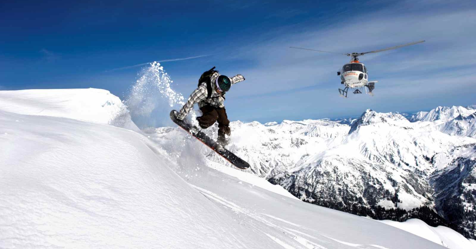 Snowboarder springt eine verschneite Bergkante hinunter, während ein Helikopter im Hintergrund über den Alpen schwebt.