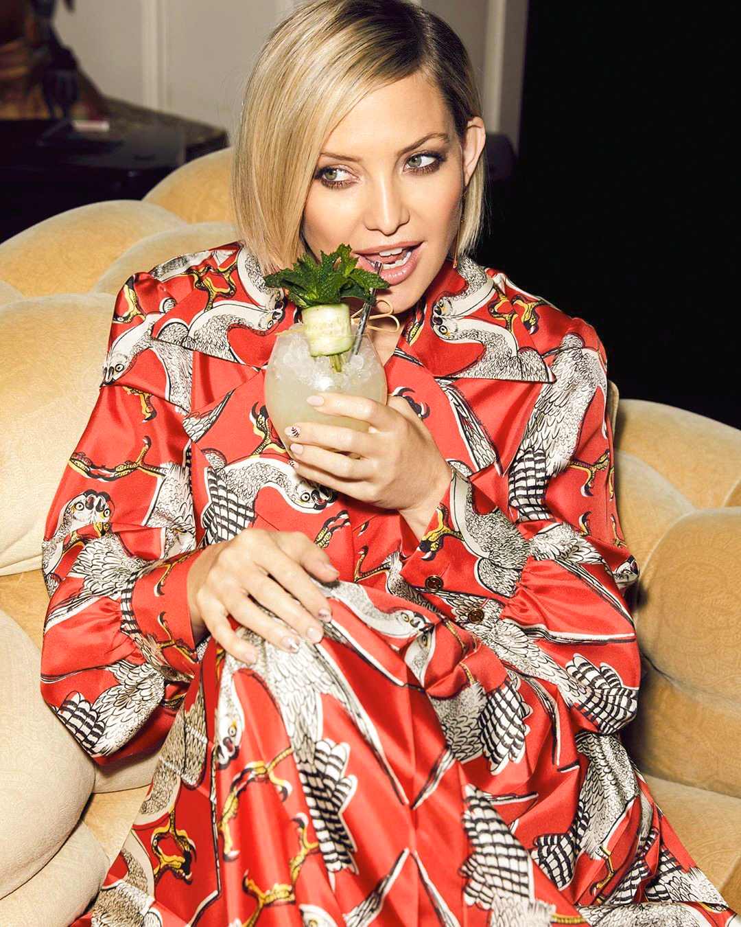 Eine Frau in einem roten, gemusterten Kleid sitzt auf einem Sofa und hält ein Cocktailglas mit Eis, Kräutern und einer Gurkenspirale.