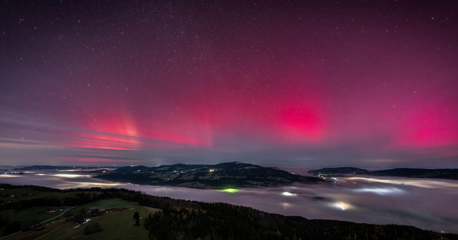 Polarlichter in kräftigen Rot- und Magentatönen über einer hügeligen Landschaft mit Nebel und verstreuten Lichtern, aufgenommen in der Nacht auf den 12. November in Niederösterreich.