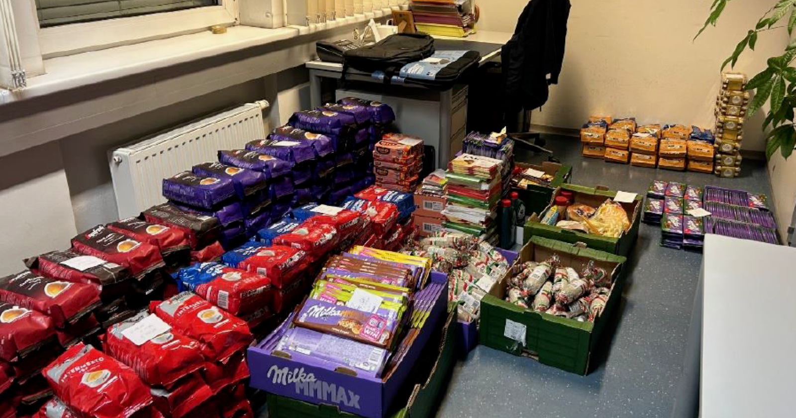 Mehrere Stapel von beschlagnahmten Lebensmitteln, darunter Kaffeepackungen, Milka-Schokolade und Wurstwaren, auf dem Boden eines Polizeibüros.