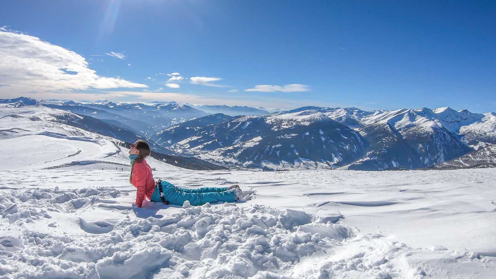 Person macht eine Yoga-Übung im Schnee auf einem Hochplateau mit weitem Blick über verschneite Berge unter klarem, blauem Himmel.
