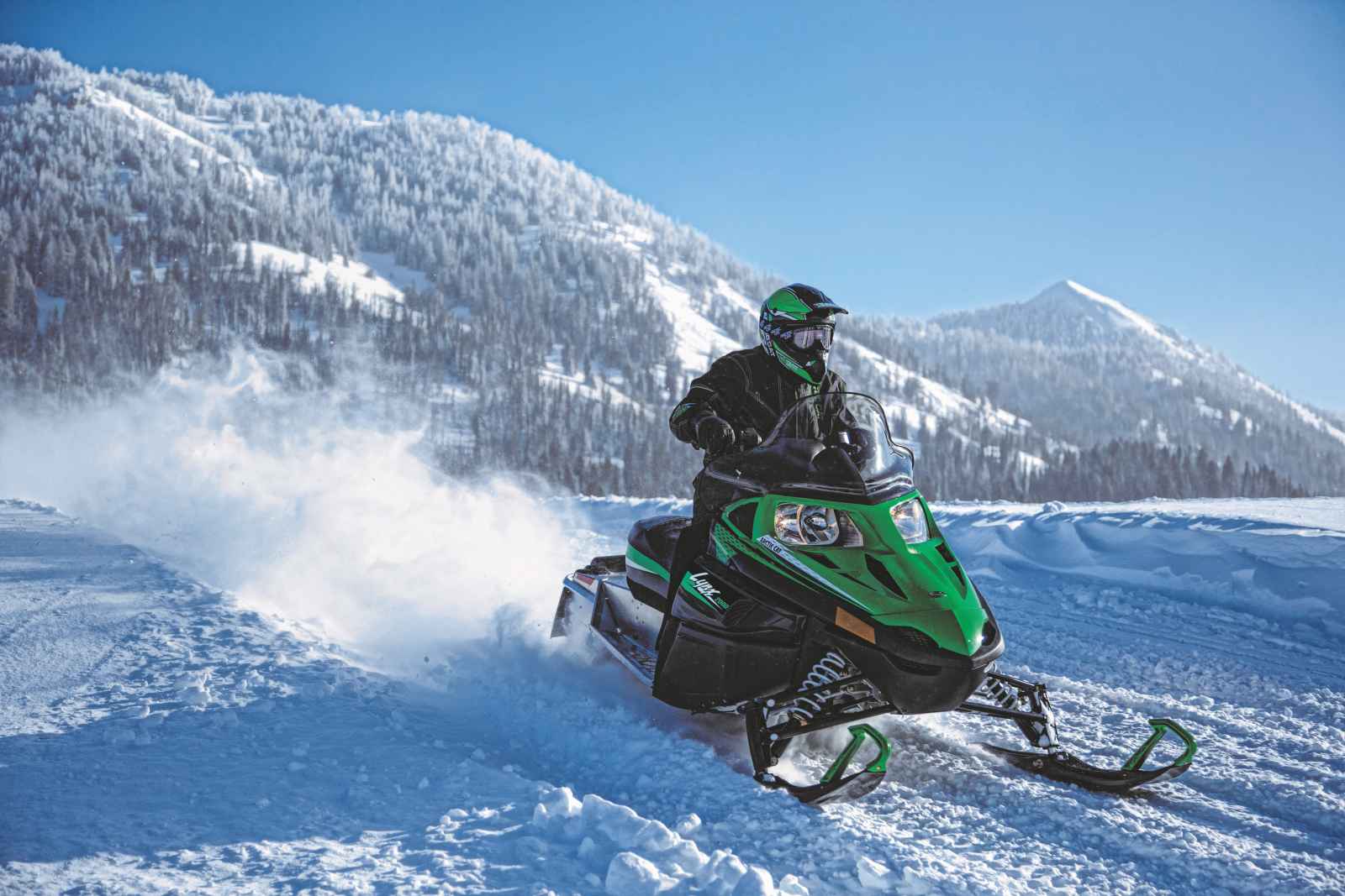 Person fährt mit einem grünen Schneemobil über eine verschneite Piste, während im Hintergrund schneebedeckte Berge und ein klarer blauer Himmel zu sehen sind.