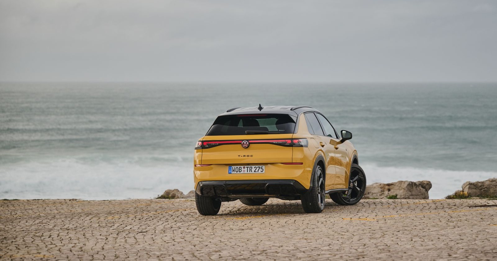 Ein gelber VW T-Roc parkt auf einem steinernen Parkplatz. Im Hintergrund ist ein Meer - mit niedrigen Wellen - zu sehen.