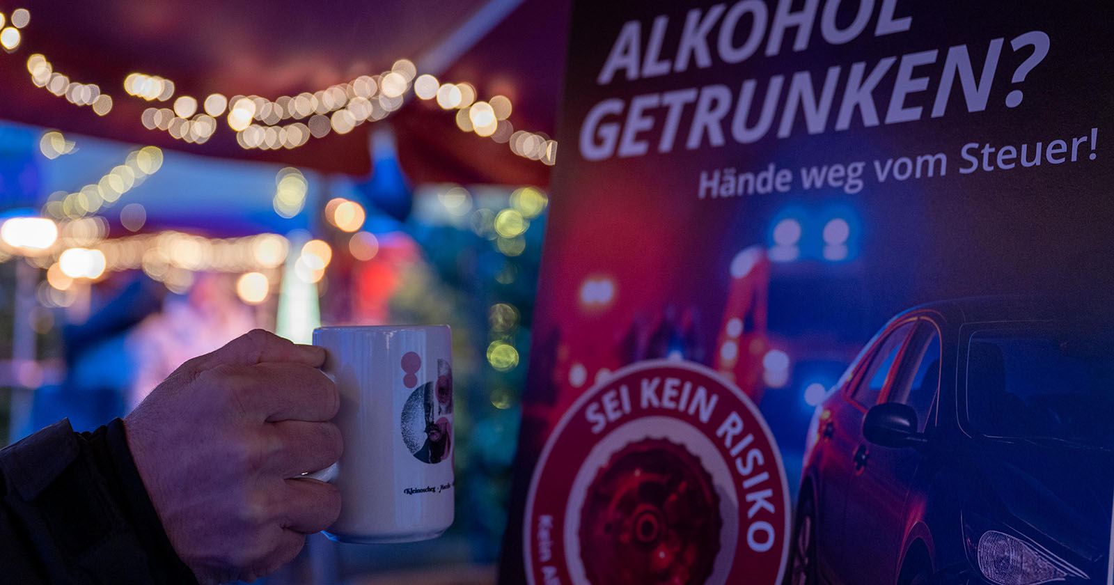 Hand die einen Punschbecher hält. Im Hintergrund ein Plakat der Aktion GEMEINSAM SICHER