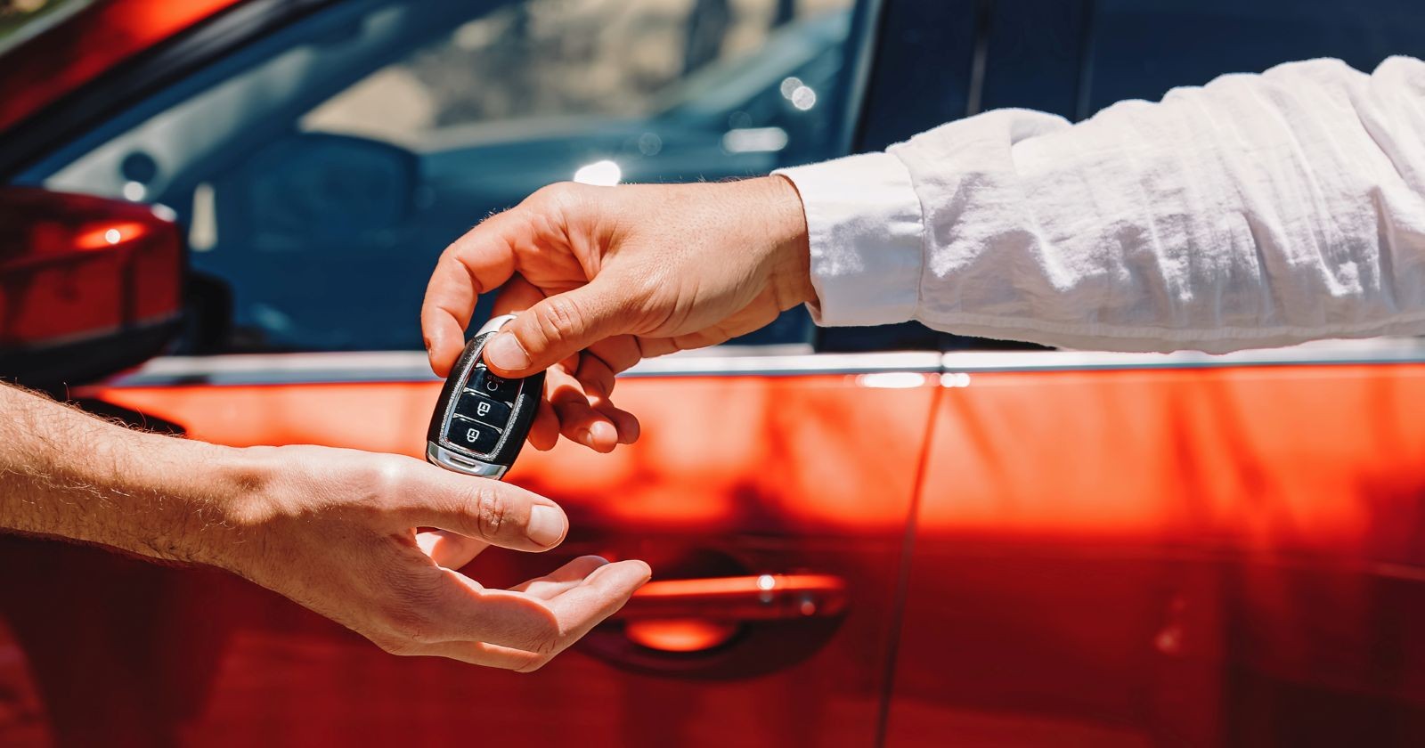 Autohändler übergibt Autoschlüssel an einen Kunden, im Hintergrund steht ein rotes Auto