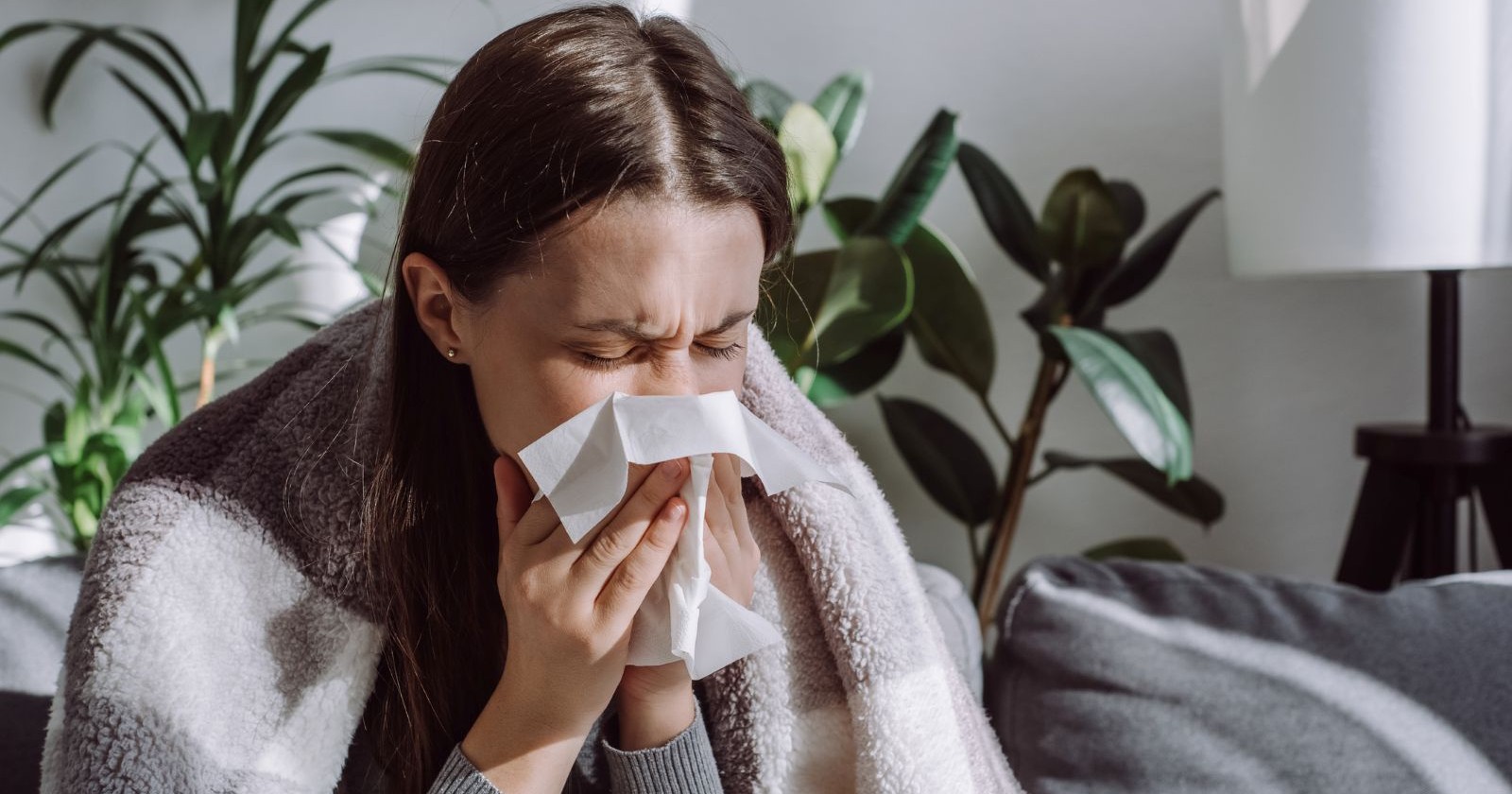 Frau mit Grippe, welche auf einem Sofa sitzt und sich die Nase putzt