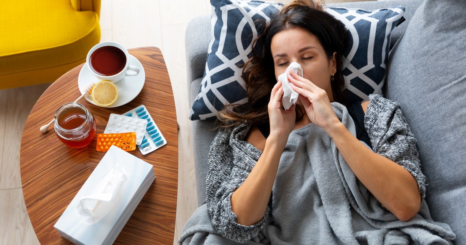 Eine Frau liegt mit Grippesymptomen auf dem Sofa, umgeben von Tee, Honig und Medikamenten – die aktuelle Influenza-Welle hat viele Menschen in Österreich erwischt, besonders in den Hotspots wie Oberösterreich