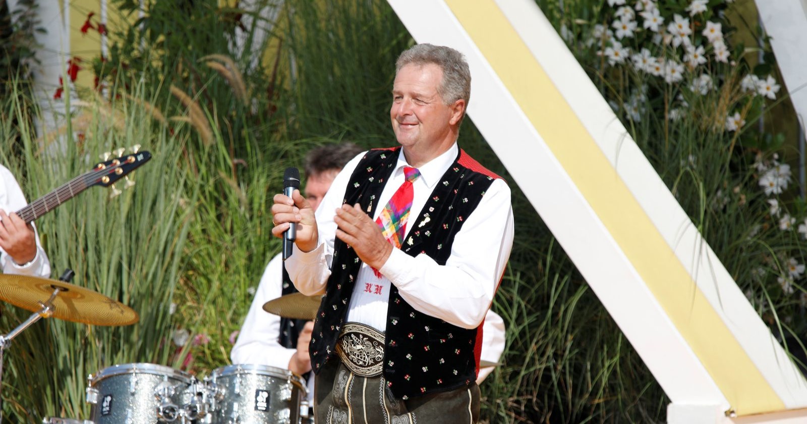Norbert Rier, Frontsänger der Kastelruther Spatzen, in Tracht hält ein Mikrofon und steht vor einer Band mit Schlagzeug und Gitarre bei einem Auftritt im Freien.