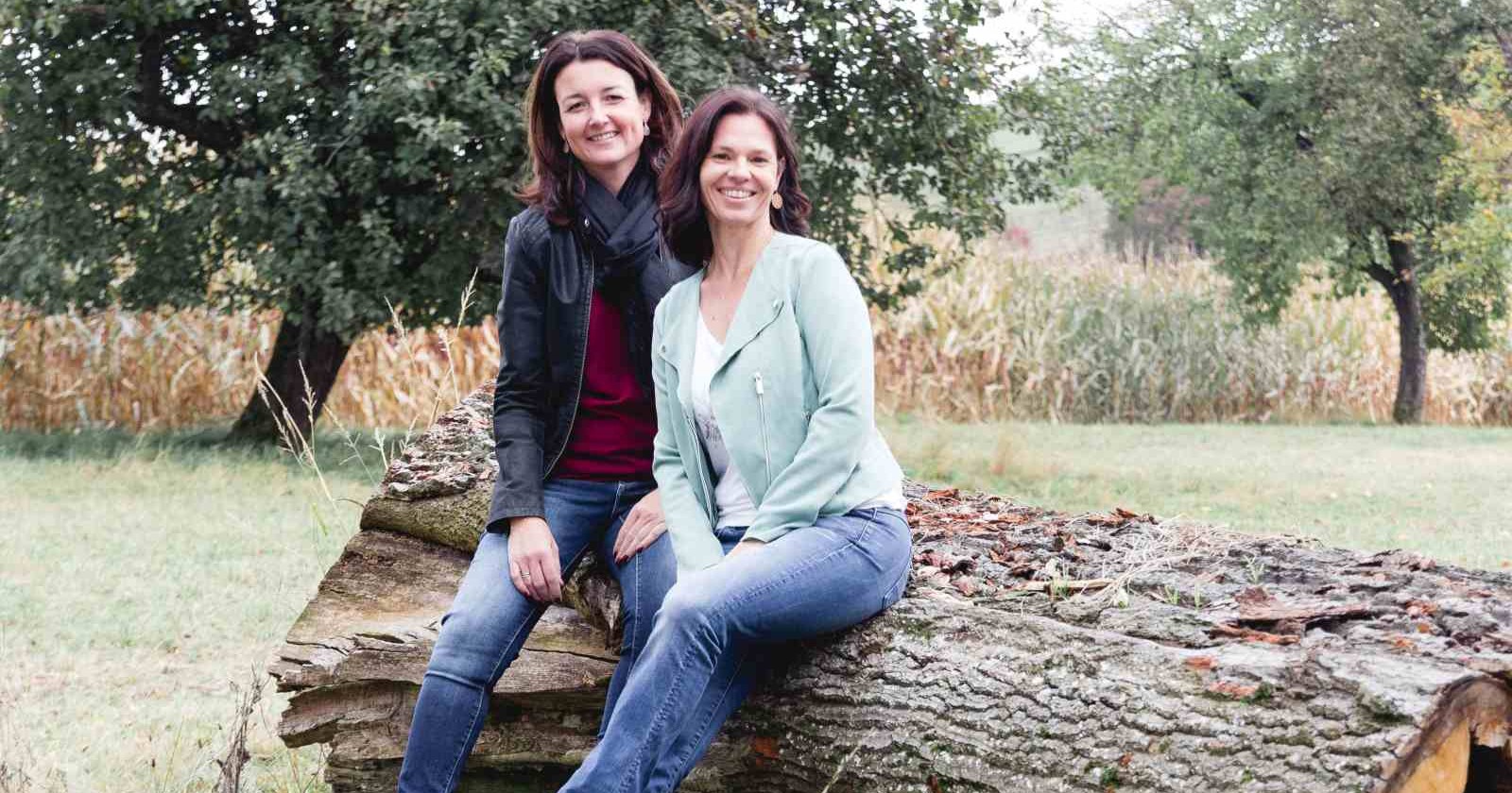 Zwei lächelnde Frauen sitzen auf einem umgestürzten Baumstamm in einer herbstlichen Wiese mit Bäumen und Maisfeld im Hintergrund.