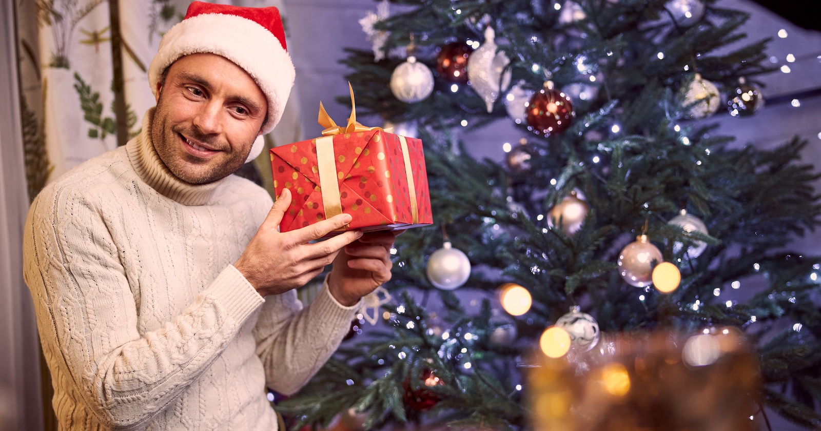 Mann mit weißem Pullover und Santa Claus-Mütze mit einem Weihnachtsgeschenk vor dem Christbaum