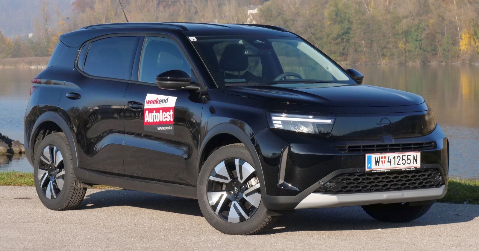 Ein schwarzer Opel Frontera parkt am Rande eine Gewässers. Das Auto leuchtet in der Sonne, im Hintergrund sind einige Bäume zu erkennen.