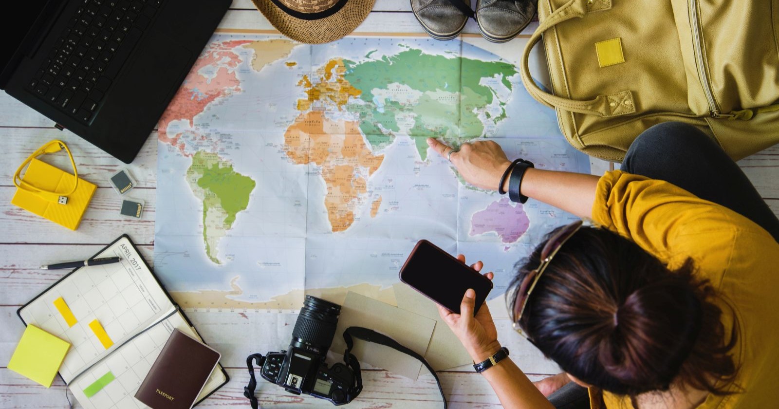 Frau zeigt auf eine Weltkarte, daneben liegen ein Pass und eine Kamera
