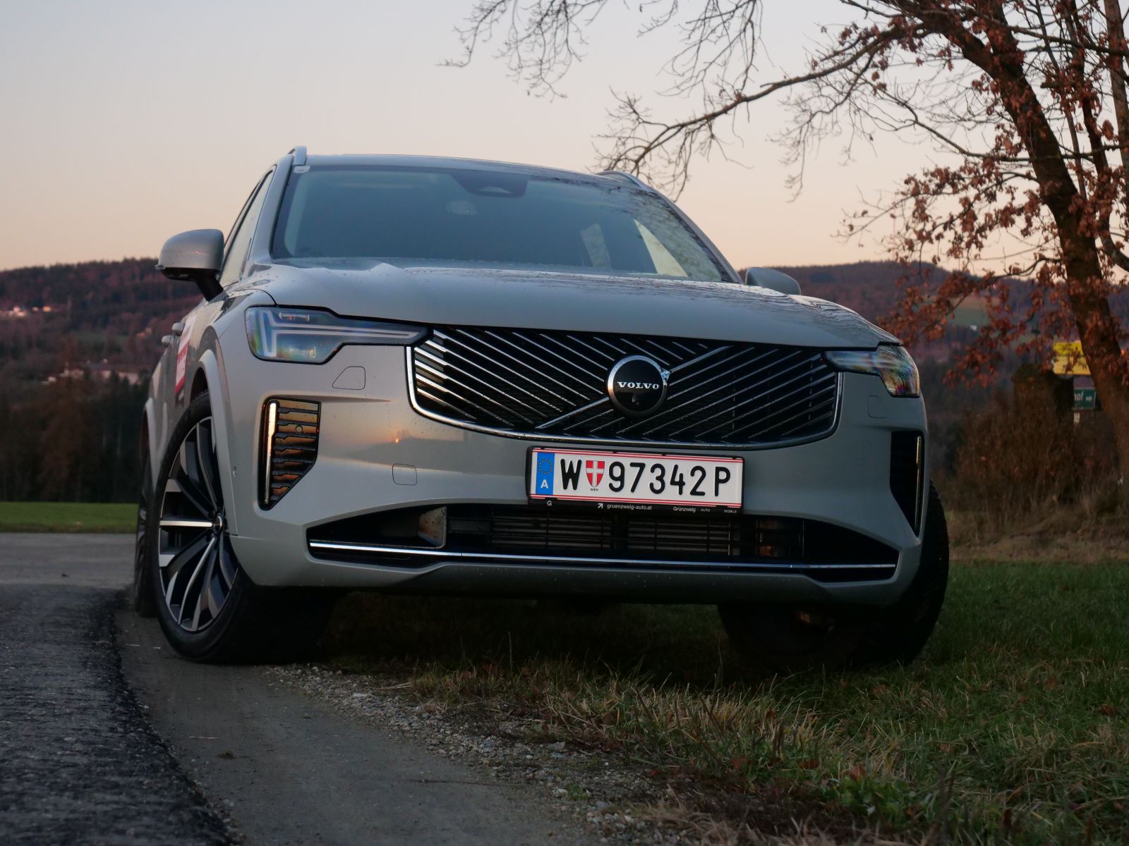 Eine silberner Volvo XC90 von vorne. Das Auto ist in der Abenddämmerung zu sehen, im Hintergrund ist eine Hügellandschaft zu erkennen.