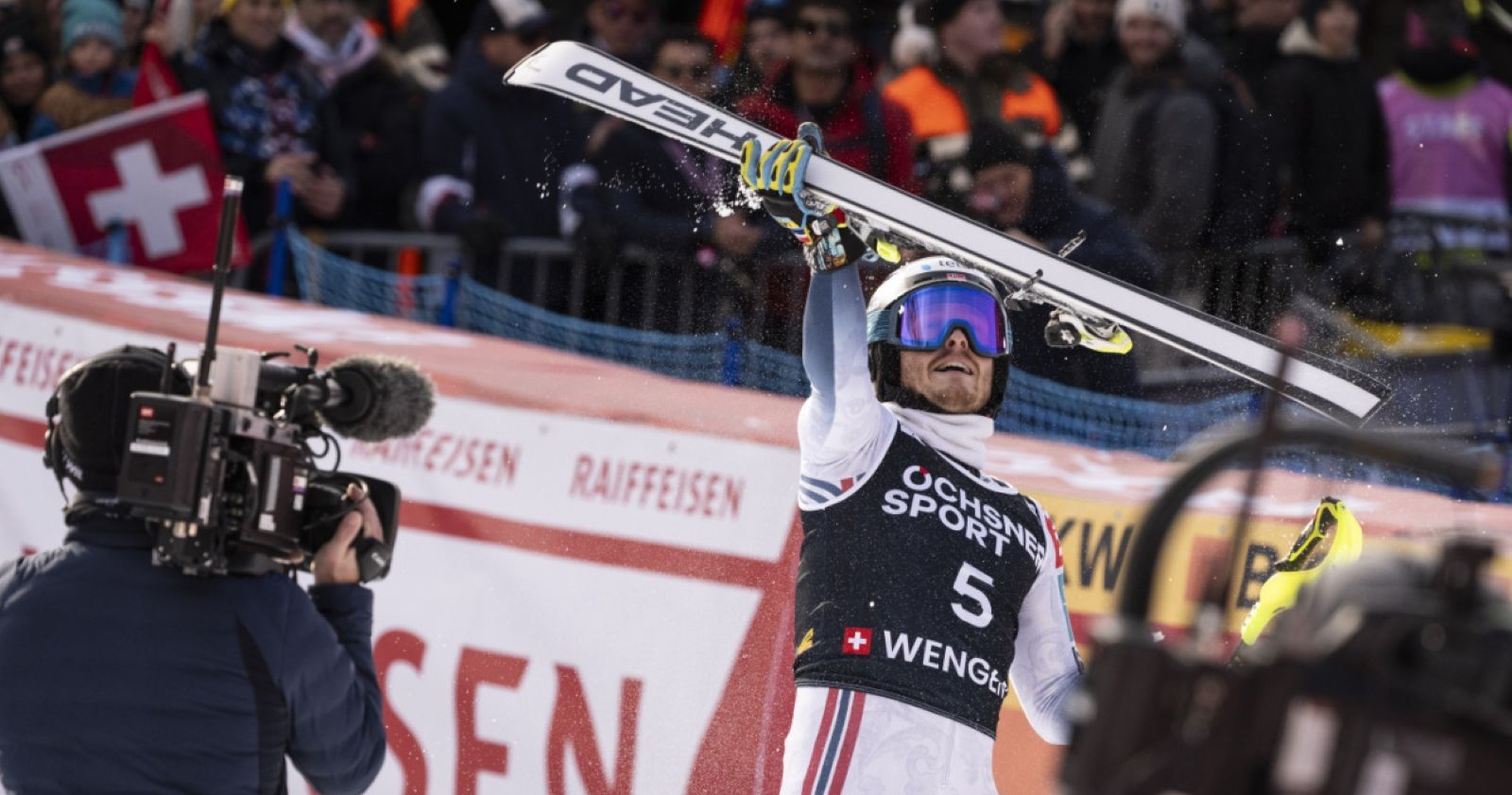 Ein Skirennläufer feiert im Zielraum von Wengen seinen Sieg, hält seine Ski triumphierend über den Kopf, während Kameras und Fans den Moment festhalten.