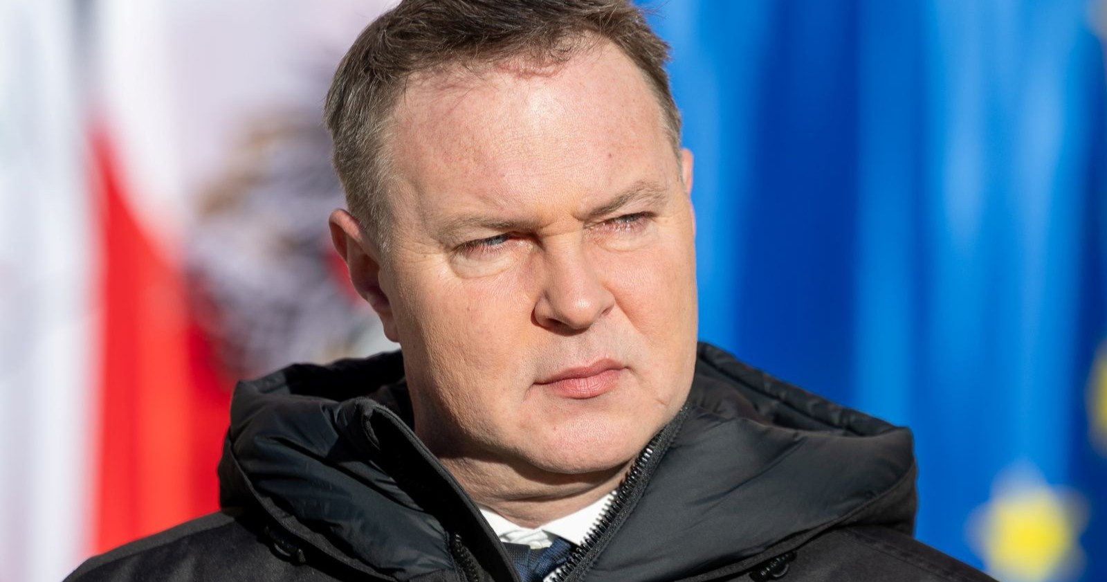 Andreas Babler blickt ernst zur Seite, im Hintergrund sind die Flaggen Österreichs und der Europäischen Union zu sehen.