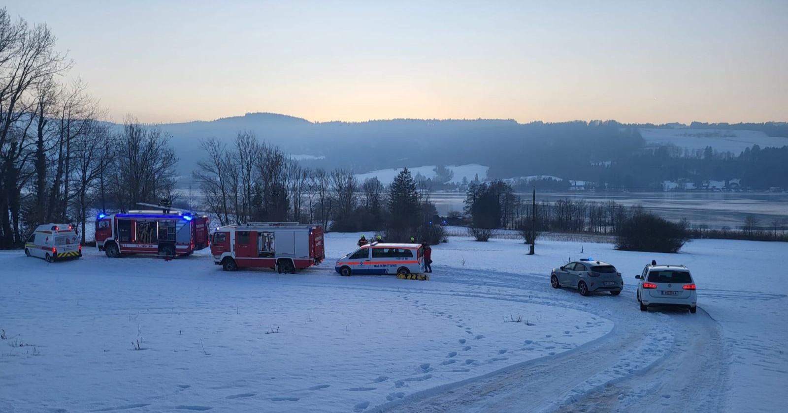 Mehrere Einsatzfahrzeuge von Feuerwehr, Wasserrettung und Polizei stehen auf einer verschneiten Wiese am Irrsee in Oberösterreich während eines Rettungseinsatzes bei winterlicher Abenddämmerung.