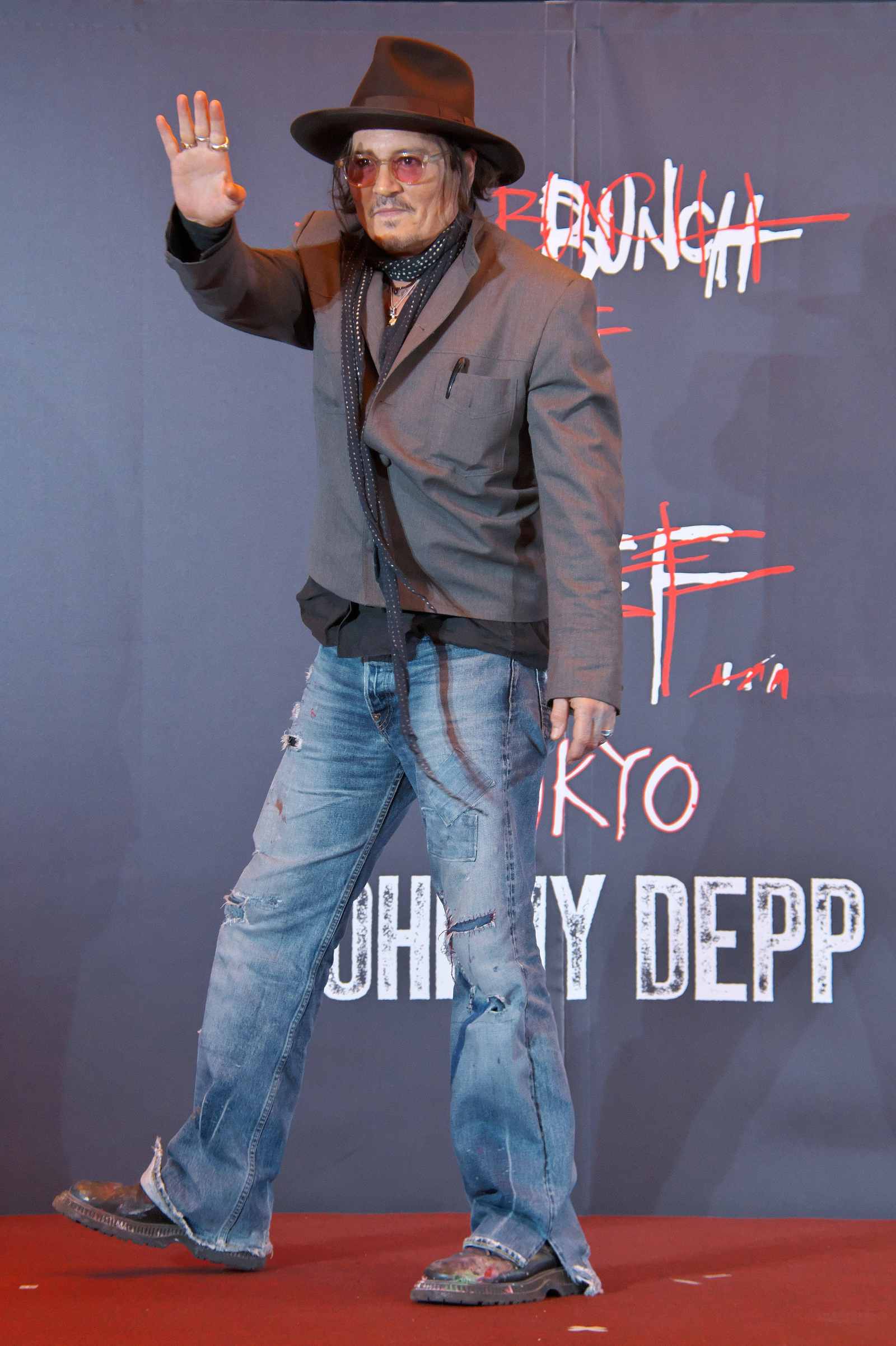 Johnny Depp winkt bei einem Event vor einer Fotowand und trägt Hut, Sonnenbrille, Sakko und Jeans.
