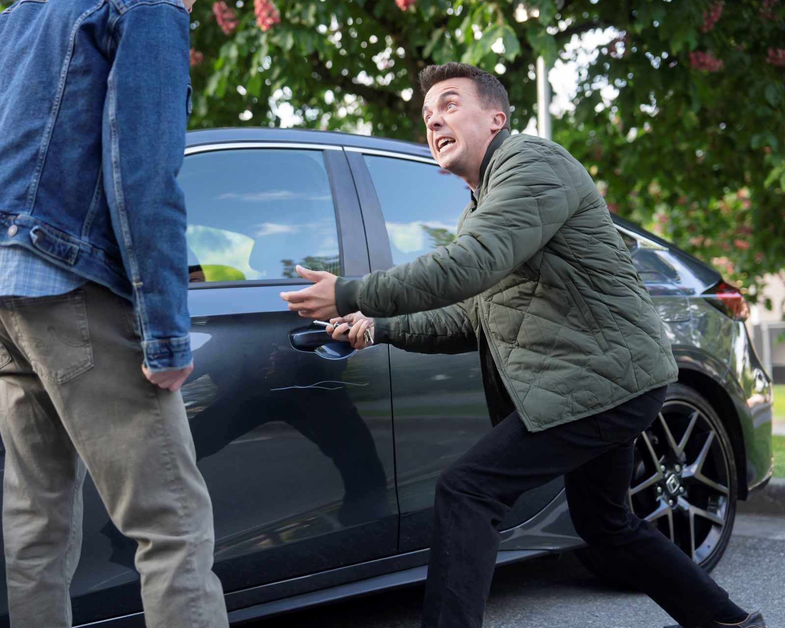 Ein Mann gestikuliert aufgebracht neben einem geparkten Auto und spricht mit einer anderen Person im Freien.