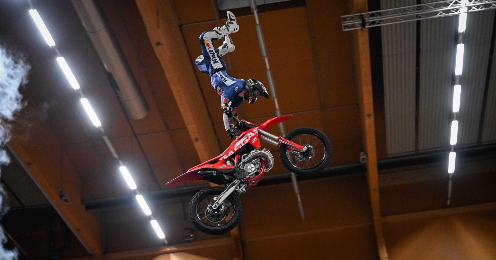 Ein rotes Motorrad fliegt durch die Luft. An den Lenker hält sich eine blau gekleideter Motorradfahrer fest. Seine Füße zeigen senkrecht nach oben, er macht einen Handstand.