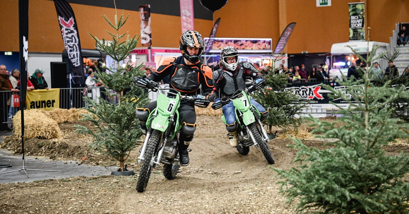 Zwei Motorradfahrer auf geländegängigen Maschinen passieren eine Strecke auf Sand. Die Strecke säumen kleine Nadelbäume, im Hintergrund sind Menschen zu sehen. Das ganze spielt sich ein einer Halle ab.