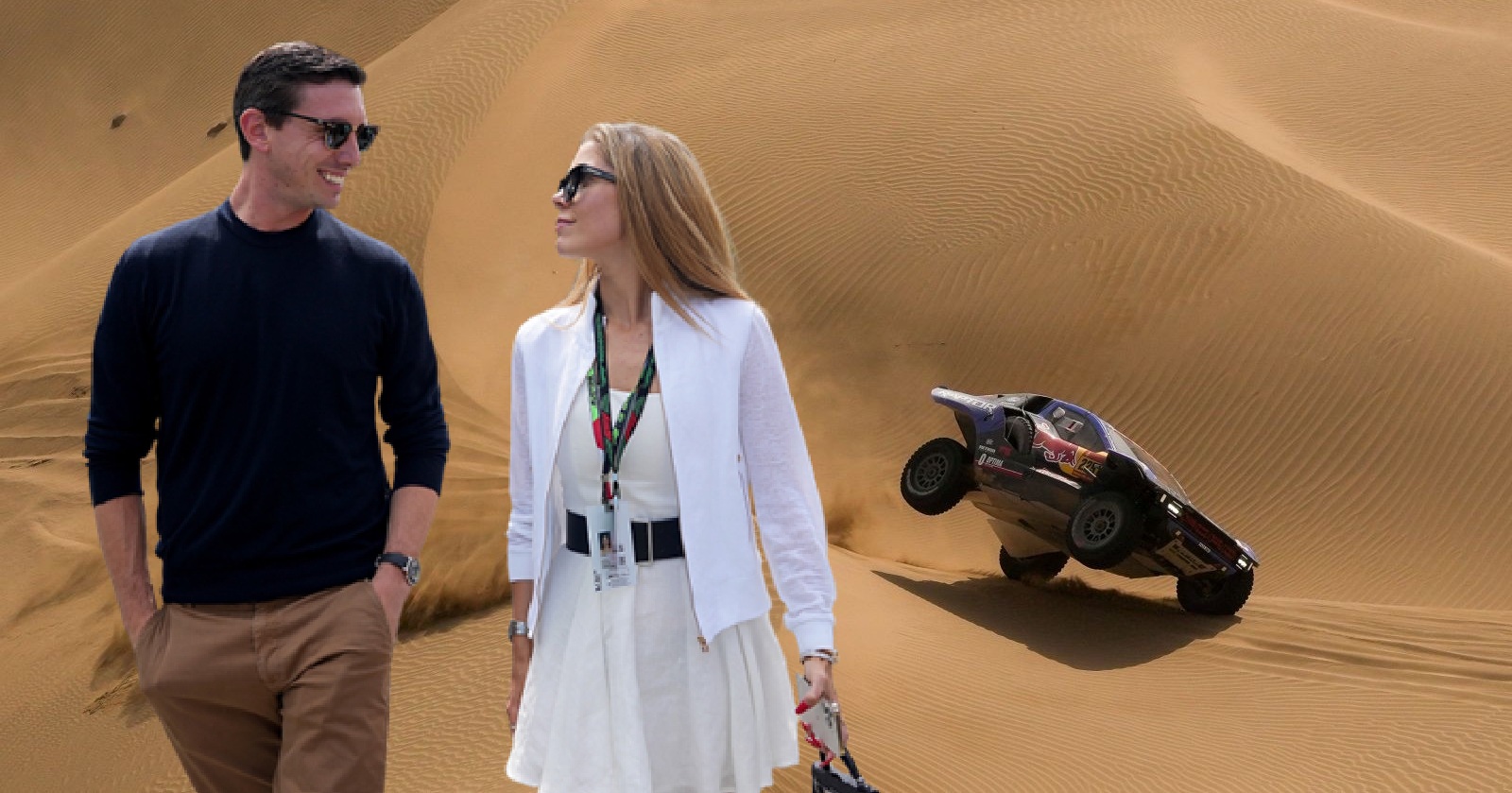 Victoria Swarovski und ihr Partner gehen lächelnd durch eine Wüstenlandschaft, während im Hintergrund ein Offroad-Fahrzeug eine Sanddüne hinauffährt.