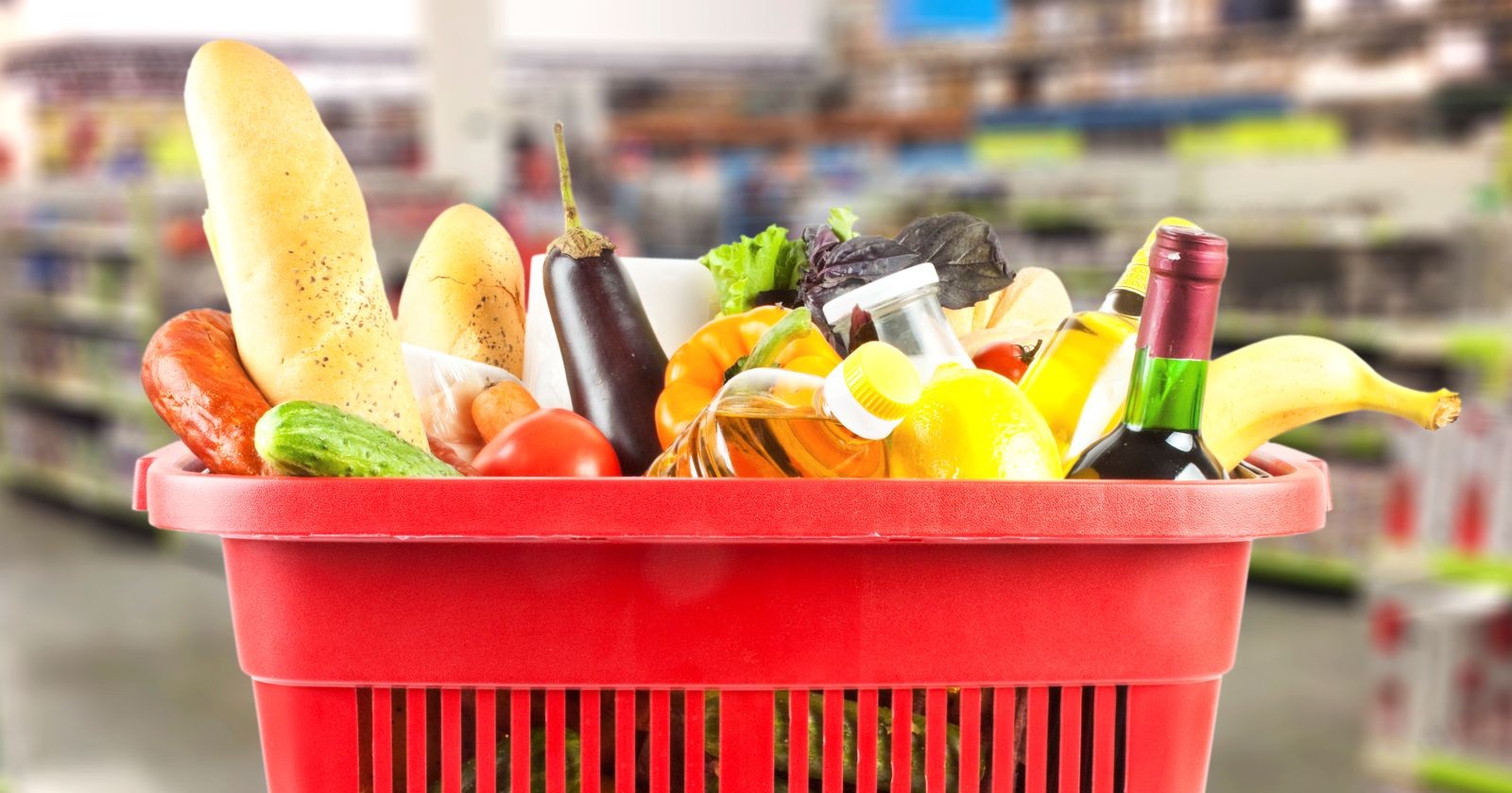 Roter Einkaufskorb mit verschiedenen Lebensmitteln wie Brot, Obst, Gemüse, Öl und Getränken in einem Supermarkt.