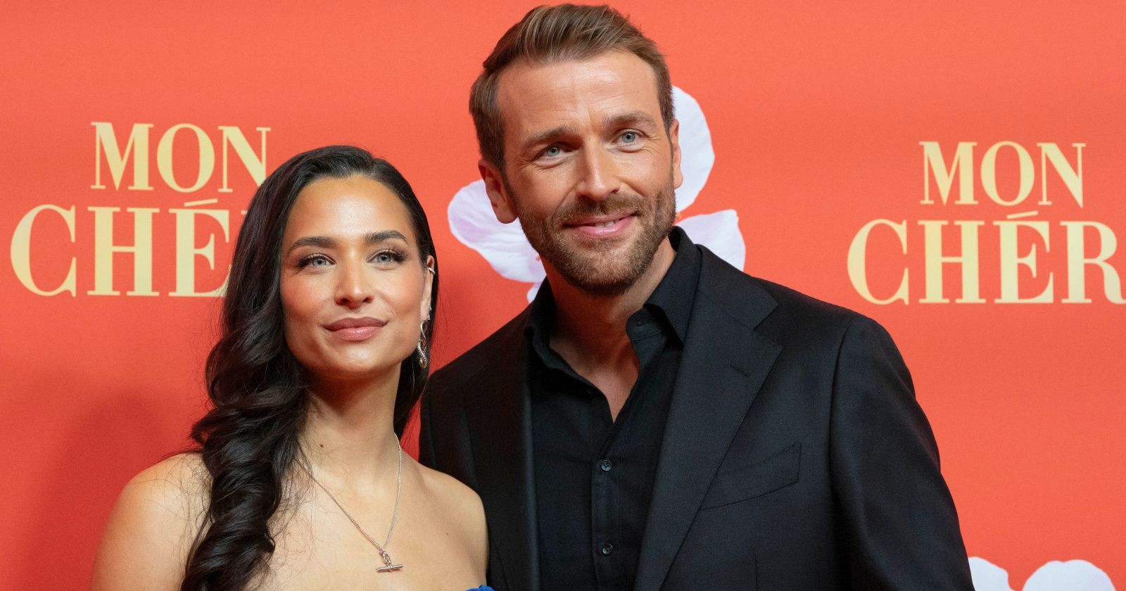 Amira Aly und Christian Düren posieren gemeinsam auf einem roten Teppich vor einer „Mon Chéri“-Fotowand und blicken lächelnd in die Kamera.