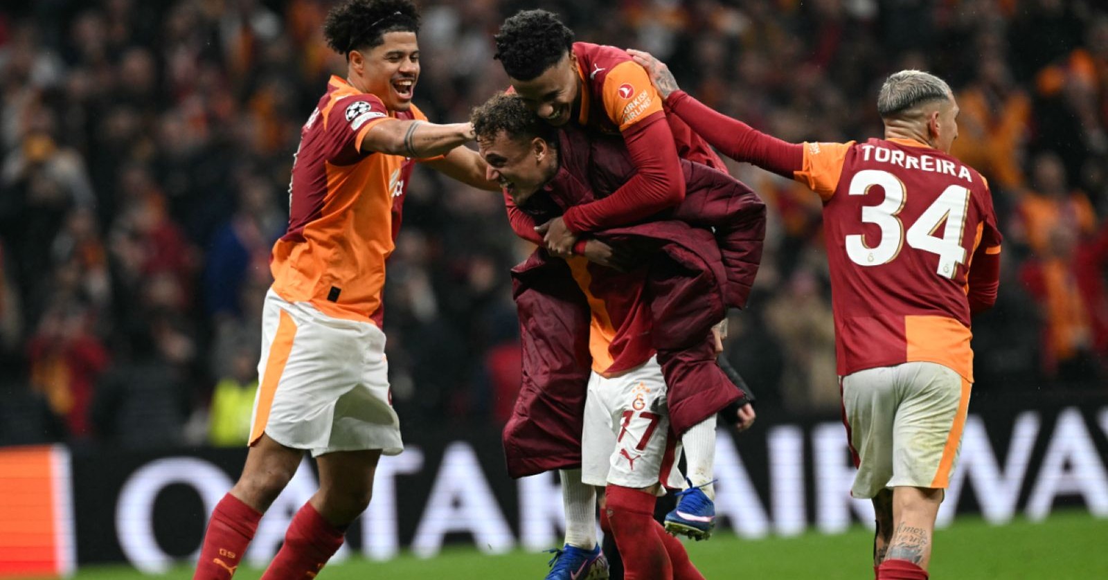 Mehrere Spieler von Galatasaray in rot-orangefarbenen Trikots jubeln gemeinsam auf dem Fußballplatz und umarmen sich nach einem Tor im Champions-League-Spiel gegen Juventus.