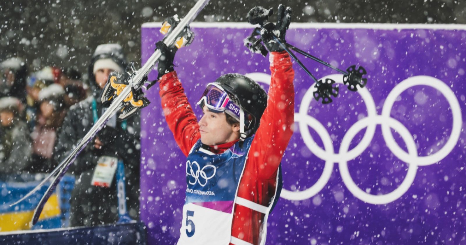 Matej Svancer hebt jubelnd seine Skier in die Luft, während Schneeflocken fallen und im Hintergrund das olympische Symbol zu sehen ist.