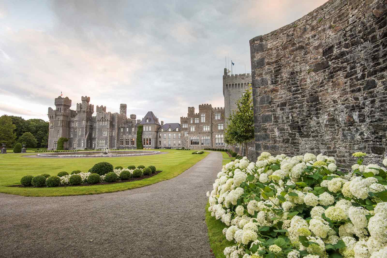 Ashford Castle in Irland mit neugotischen Türmen und Zinnen, gepflegten Rasenflächen und blühenden Hortensien entlang eines Kieswegs.