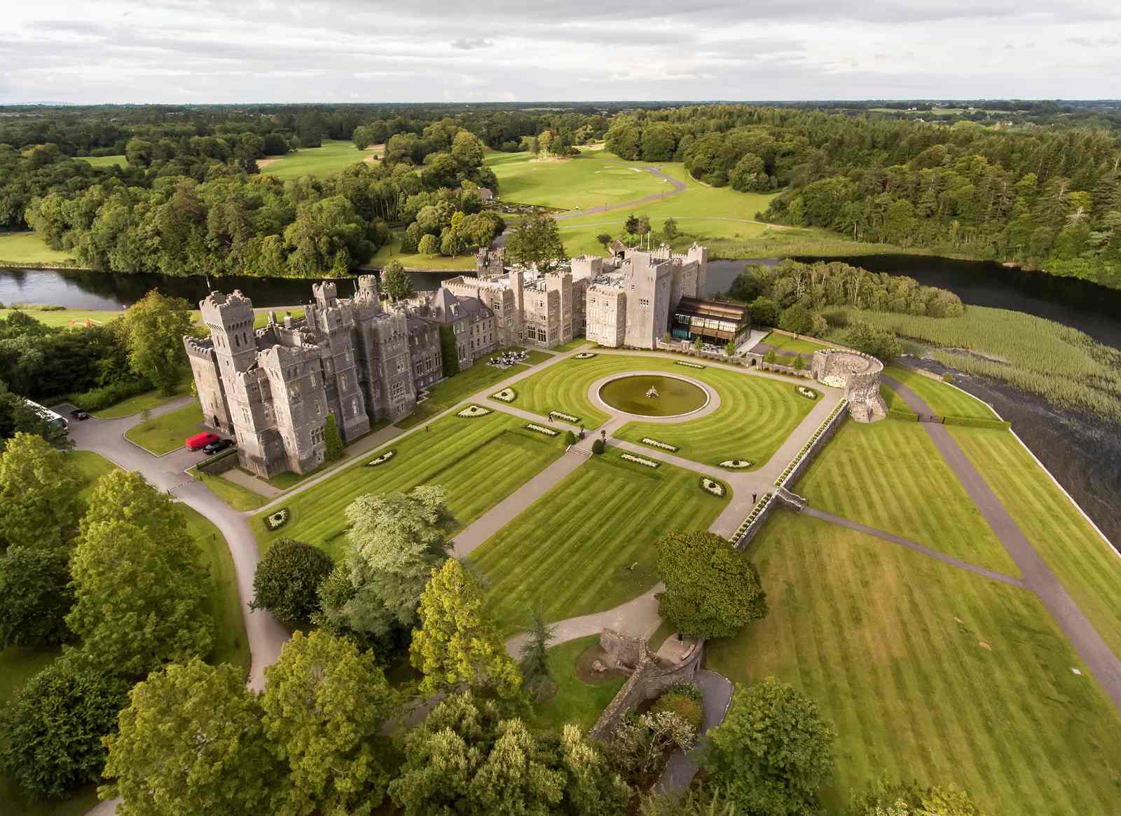 Luftaufnahme von Ashford Castle in Irland, umgeben von weitläufigen Parkanlagen, Flussläufen und grüner Landschaft.
