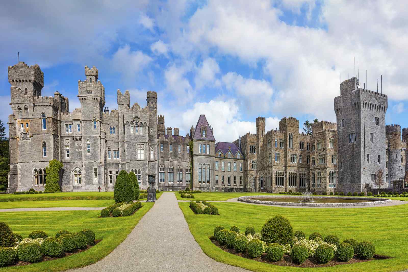 Frontansicht von Ashford Castle mit hohen Türmen, grauer Steinfassade, symmetrischem Garten und rundem Springbrunnen.
