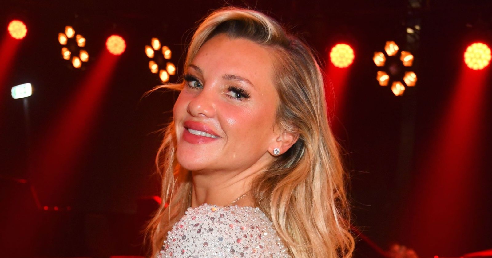 Evelyn Burdecki lächelt bei rotem Bühnenlicht bei einem Event in Berlin in einem glitzernden Kleid in die Kamera – beim Wiener Opernball 2026 lief der Auftritt nicht so glatt, sie stolperte mit der langen Schleppe ihres Kleides – Sturz auf der Treppe
