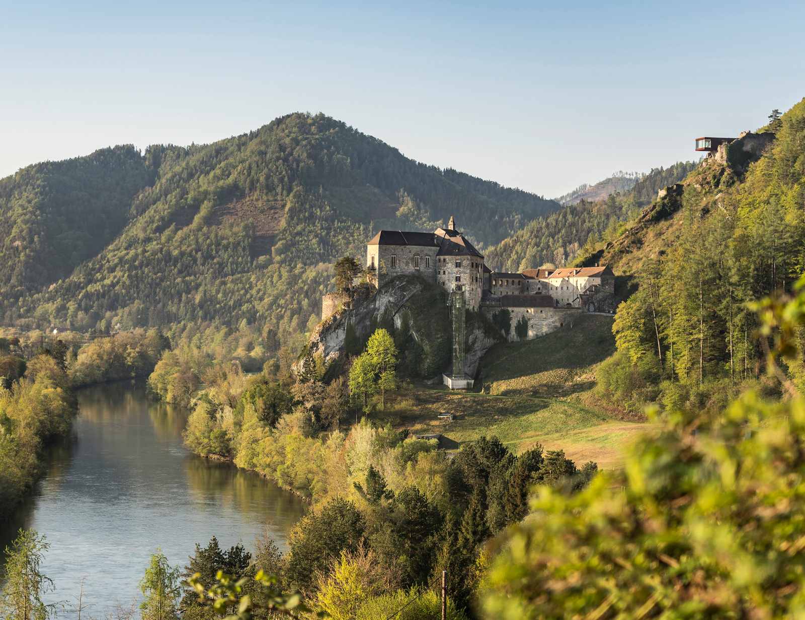 Burg Rabenstein in der Steiermark thront auf einem Felsen über der Mur, umgeben von bewaldeten Hügeln und Flusslandschaft.