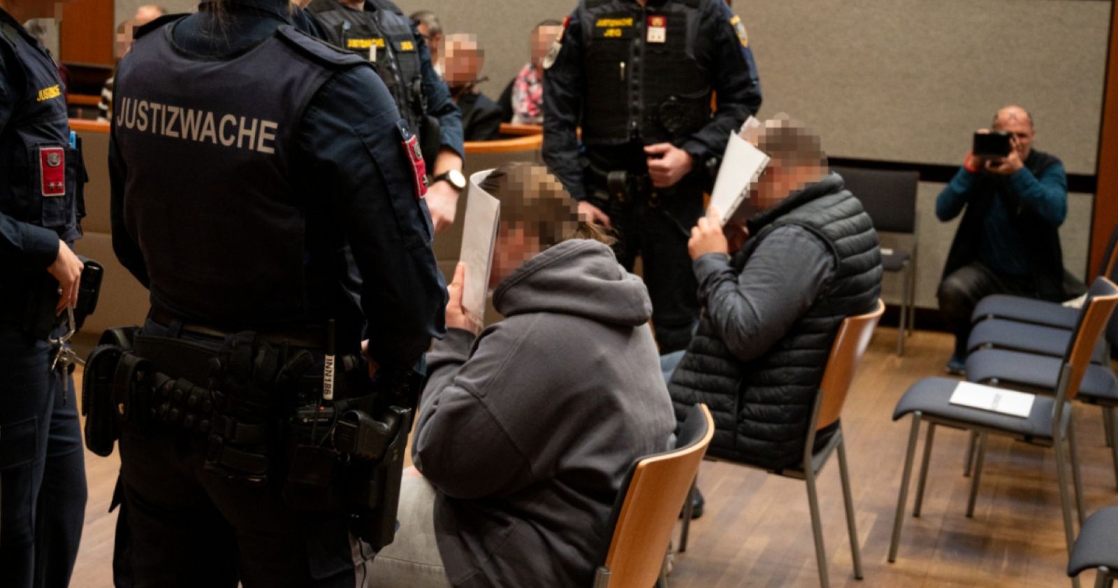 Angeklagte Eltern sitzen beim Prozess in Innsbruck auf Stühlen, verdecken ihre Gesichter mit Papierblättern, umgeben von Justizwachebeamten.