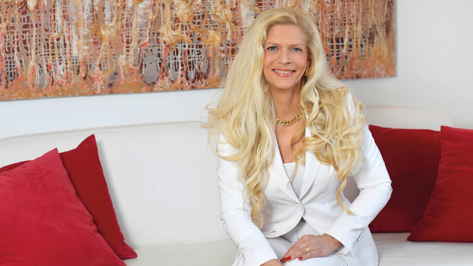 Evelyn Hendrich, sitzend auf einem hellen Sofa, mit weißem Hosenanzug und langen blonden Haaren, vor einem abstrakten Kunstwerk.