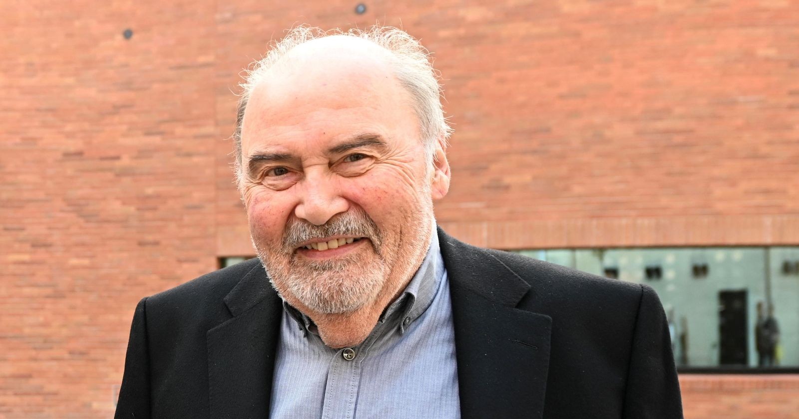 Lambert Hamel lächelt in die Kamera, trägt ein Sakko und ein Hemd; im Hintergrund ist eine Backsteinwand zu sehen.