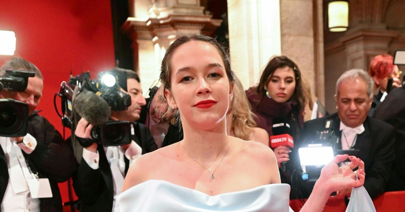 Lena Schilling in einem weißen Kleid beim Wiener Opernball, umgeben von Kameras und Journalisten, während sie mit einer politischen Botschaft protestiert.