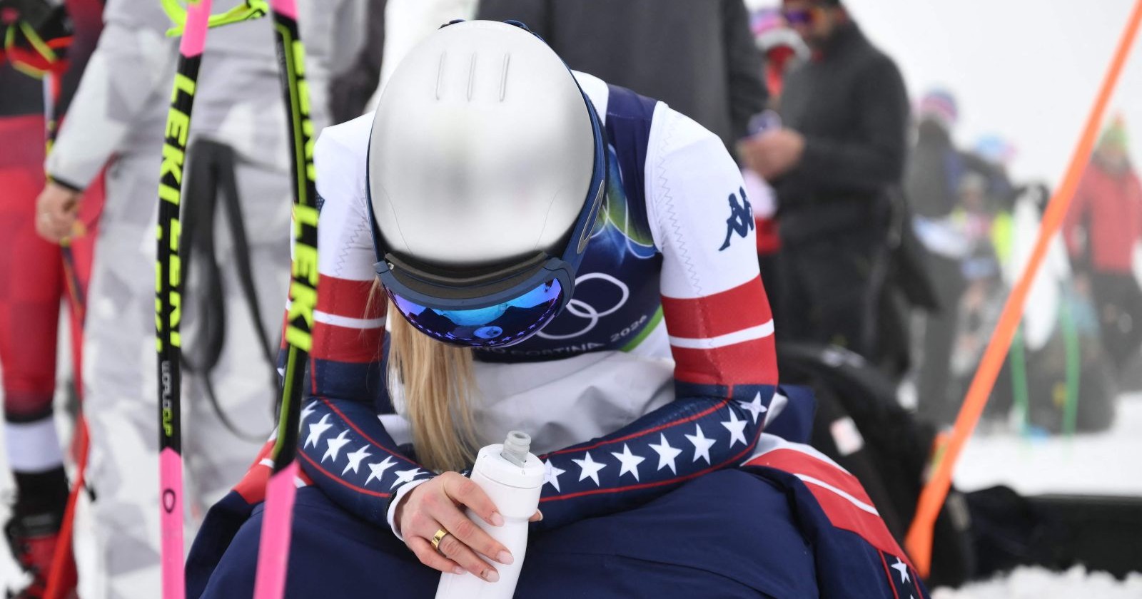US-Skirennläuferin Lindsey Vonn im Olympia-Rennanzug sitzt erschöpft im Schnee, während das Abfahrtstraining in Cortina wegen Nebel und schwieriger Bedingungen gestoppt ist.