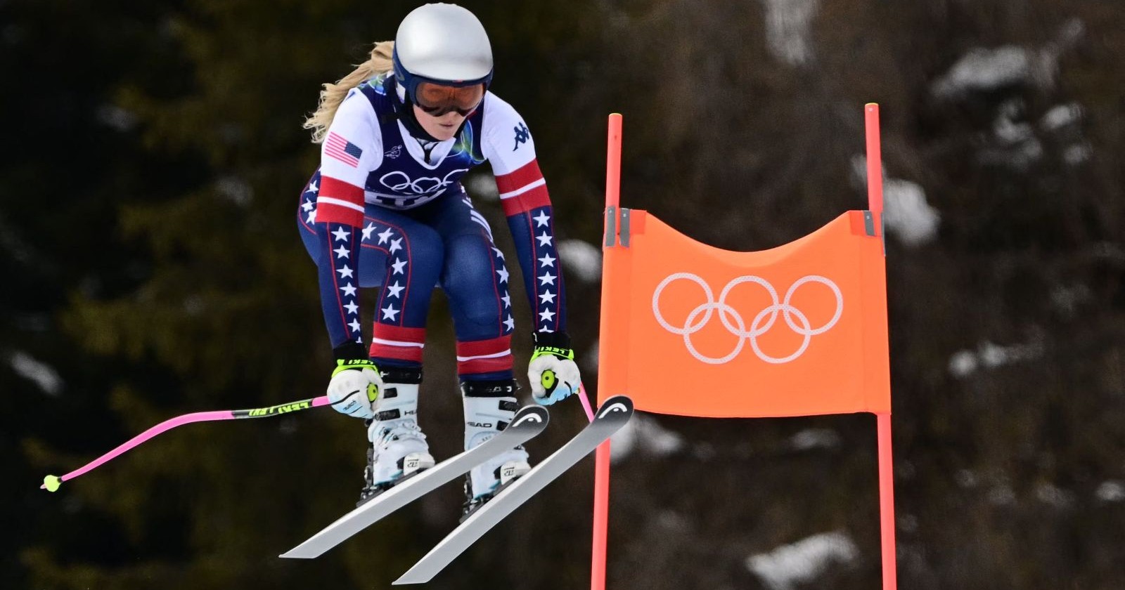 Lindsey Vonn springt im olympischen Abfahrtstraining in Cortina d’Ampezzo über ein Tor mit Olympischen Ringen, trägt Rennanzug und Helm bei schwierigen Wetterbedingungen