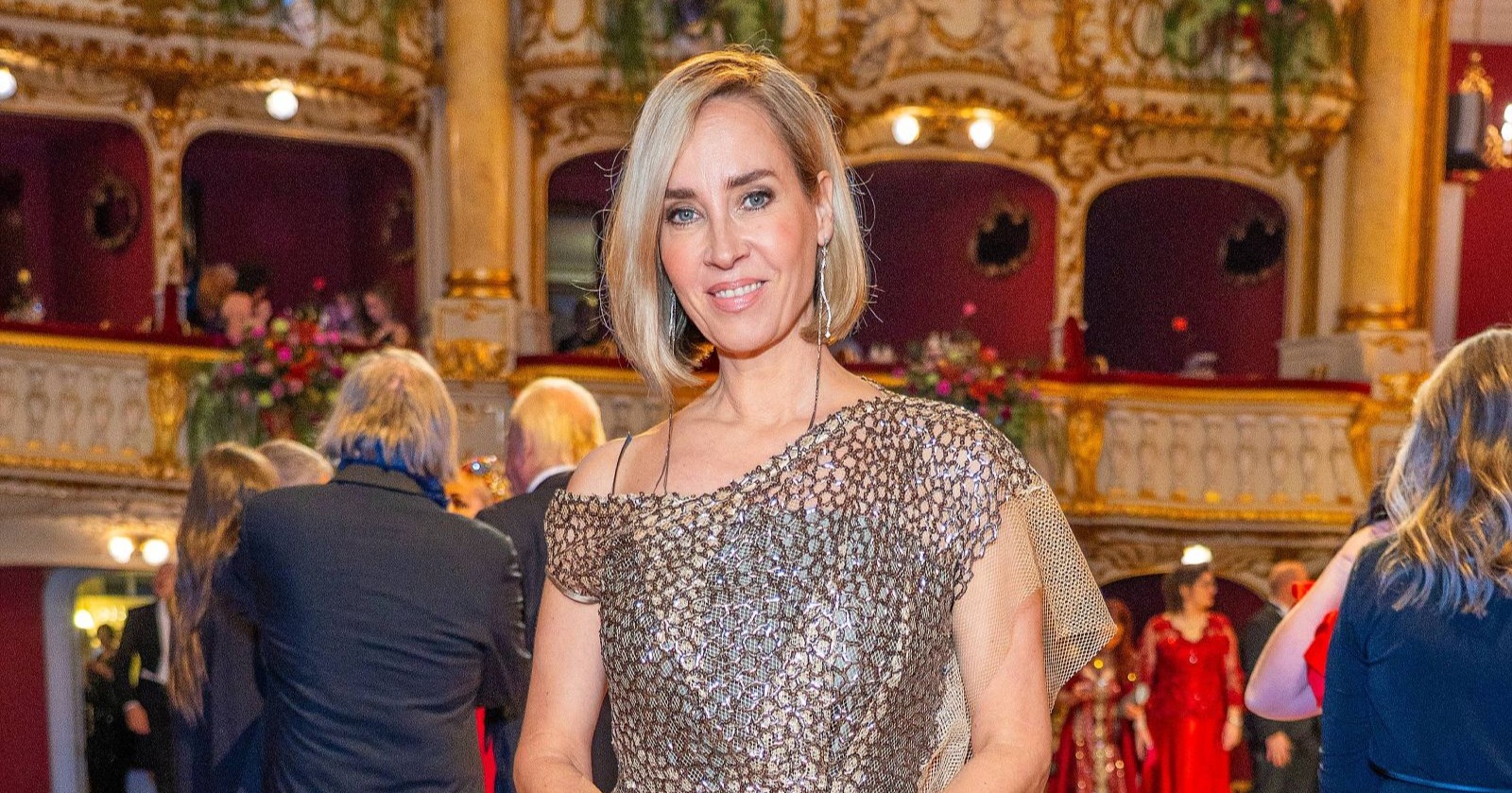 Nadja Bernhard in einem goldfarbenen Abendkleid in einem prunkvollen Theatersaal mit roten Logen im Hintergrund.