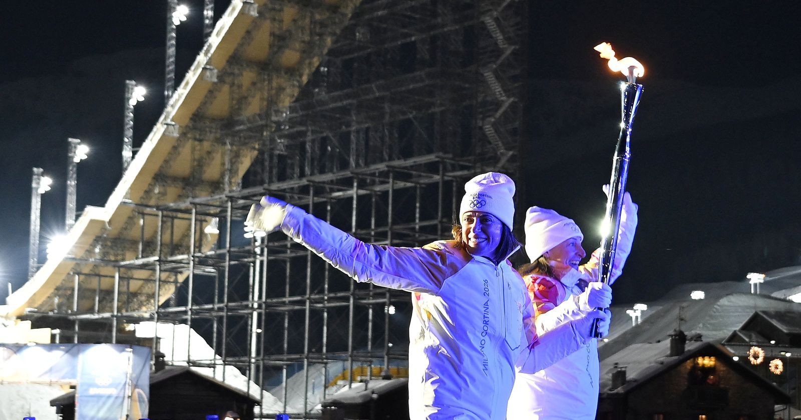 Zwei Flammenträgerinnen in weißen Olympia-Jacken tragen bei Nacht das olympische Feuer, im Hintergrund eine beleuchtete Skisprungschanze in Cortina d’Ampezzo./ bei den Olympischen Spielen 2026 werden zwei Feuer gezündet