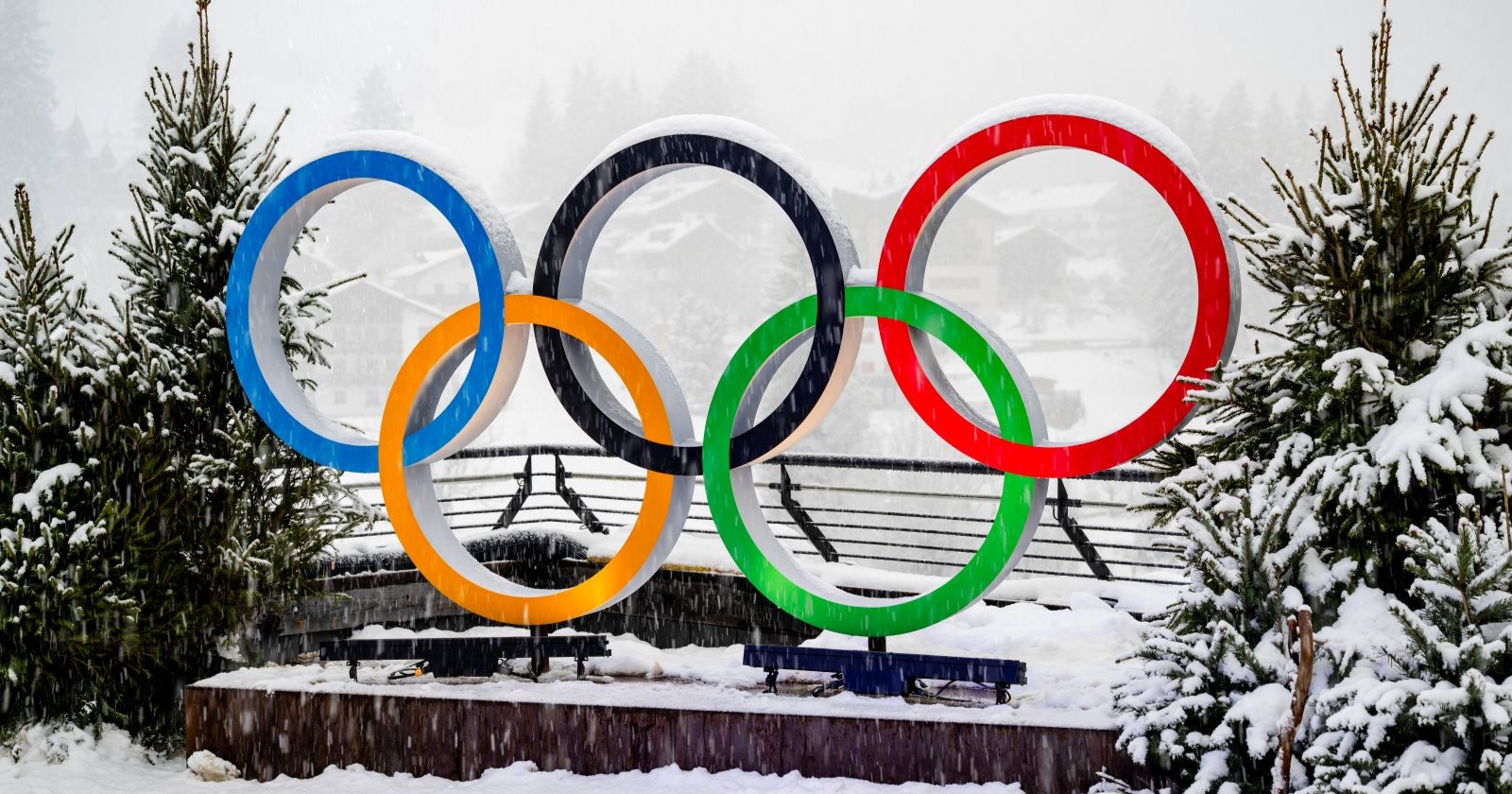 Fünf farbige Olympische Ringe stehen verschneit im Freien zwischen Tannenbäumen – winterliche Szenerie als Symbol der Olympischen Winterspiele 2026.