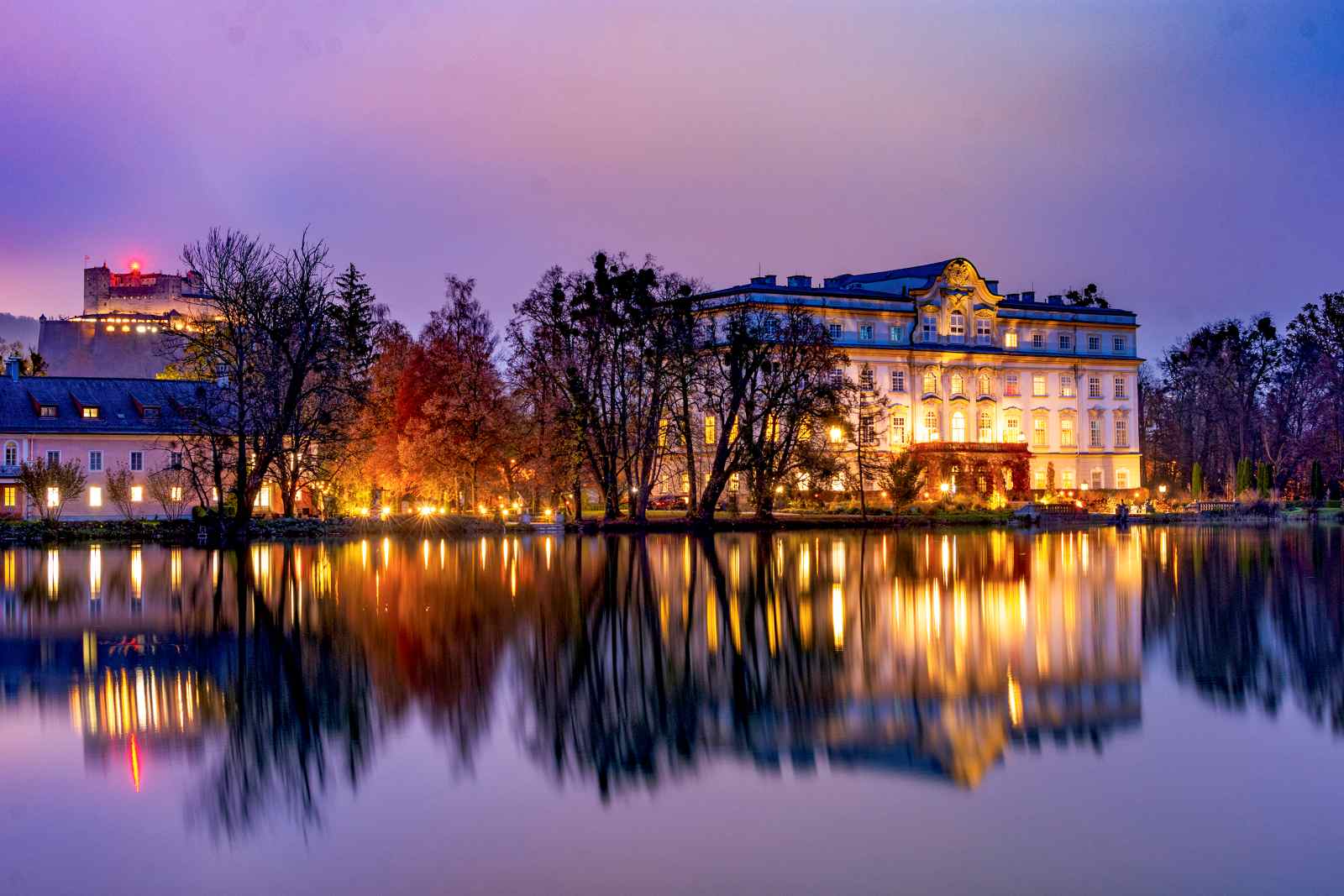 Barockes Schloss Leopoldskron in Salzburg, beleuchtet in der Abenddämmerung, mit Spiegelung im Weiher und der Festung Hohensalzburg im Hintergrund.