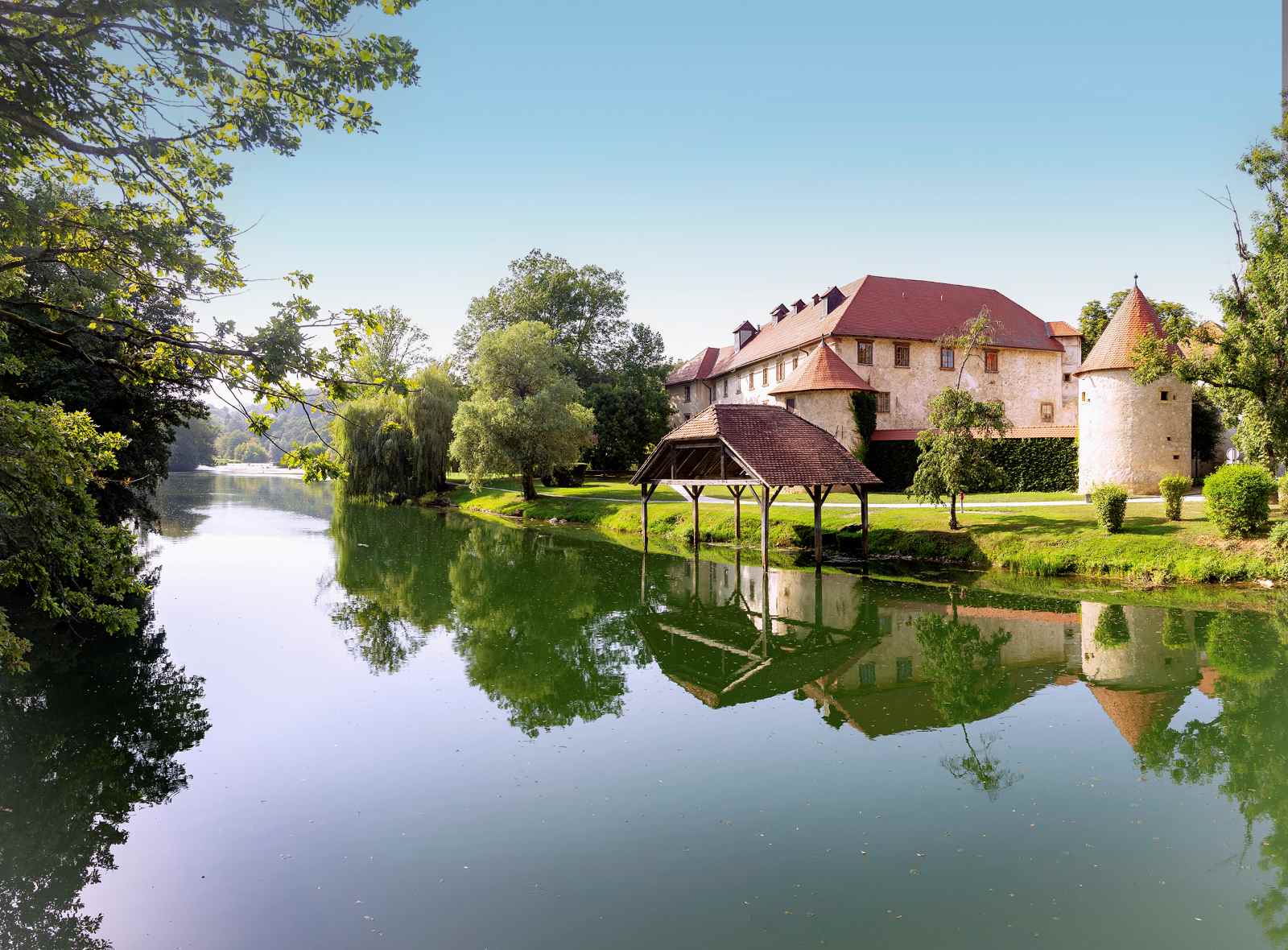 Schloss Otočec in Slowenien, ein mittelalterliches Wasserschloss auf einer kleinen Flussinsel, umgeben von Bäumen und spiegelndem Wasser.